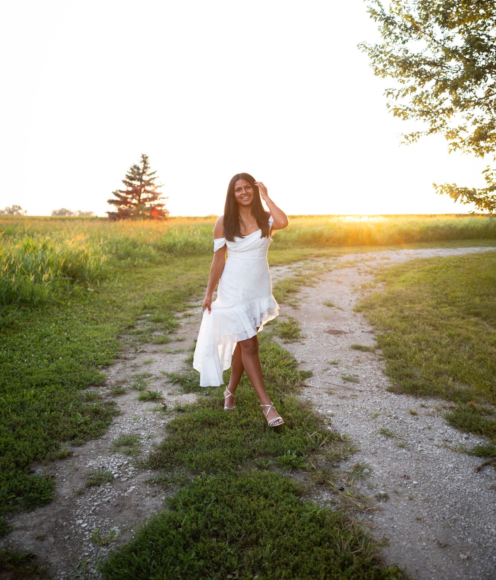 senior girl in white dress walking down lane at sunset