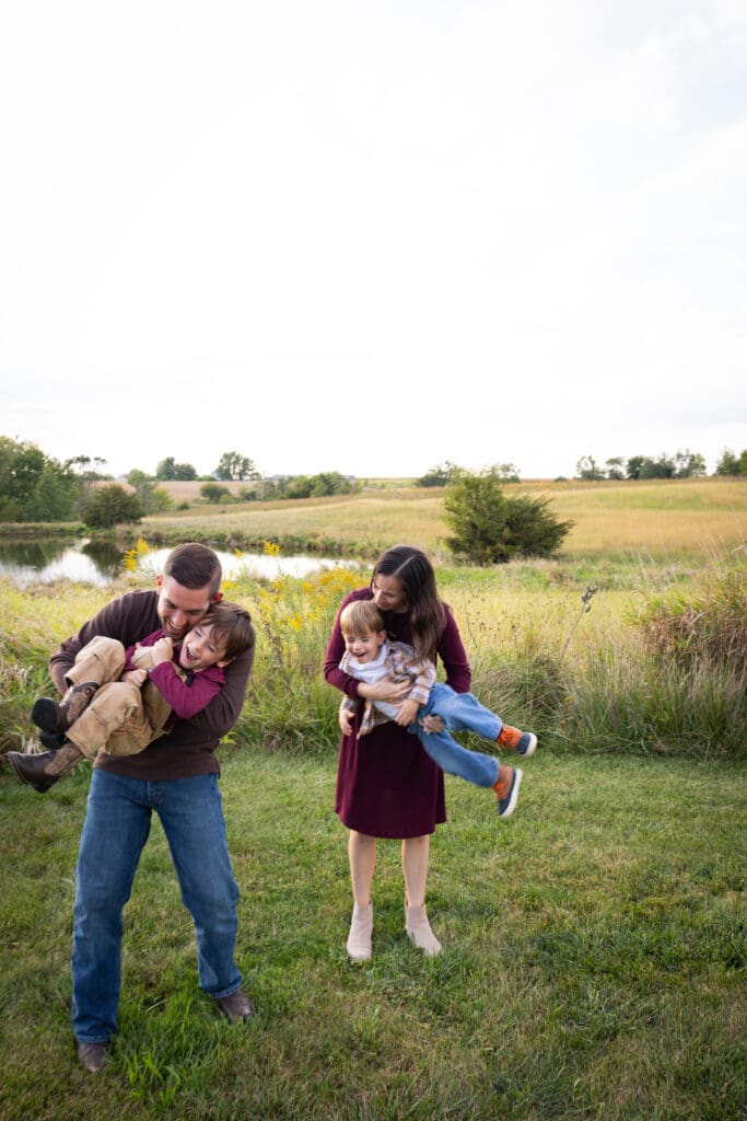 mom and dad playing with kids near tall grass and pond