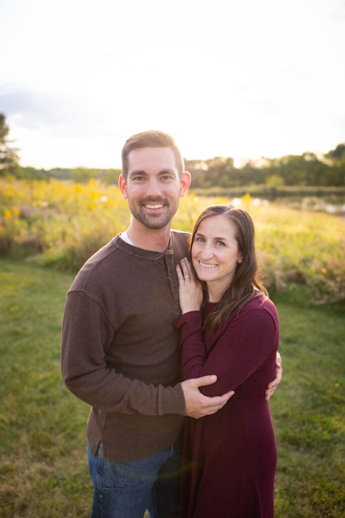 Couple smiling and embracing in a field with sunset light behind them.