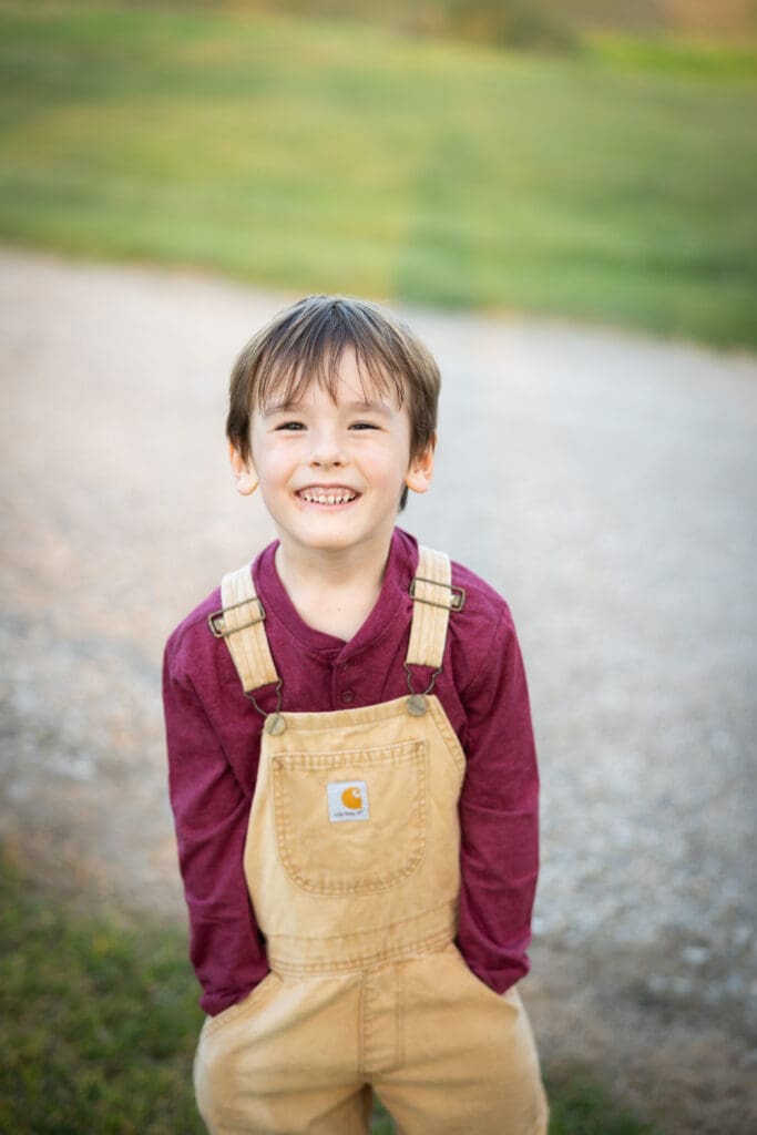 Young boy standing on gravel with hands in pockets, smiling.